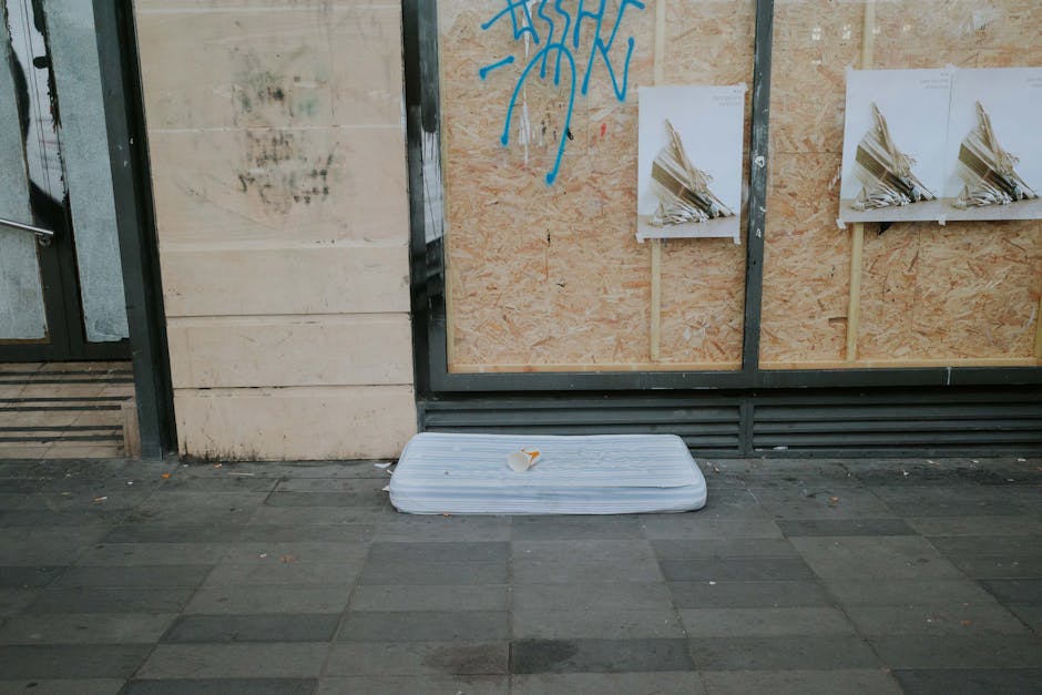 A discarded white foam mattress pad with a small paper cup resting on top, placed on a paved sidewalk in front of a building wall. The wall is partially boarded up with large sheets of plywood, which 