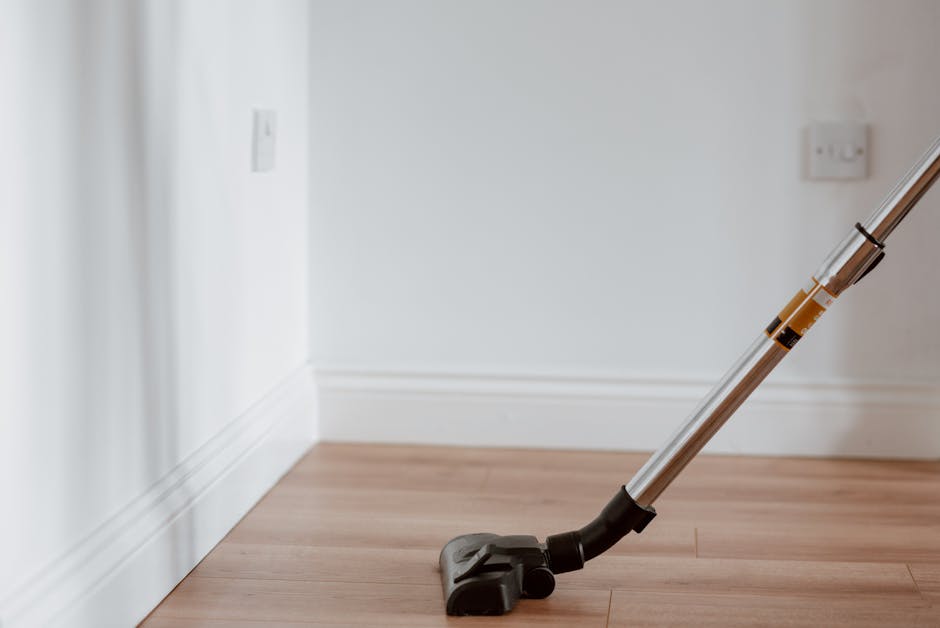 A vacuum cleaner with a silver extension tube and black floor head resting on a light wooden floor in a room with white walls and white baseboards, illustrating surface cleaning for end of tenancy sanitisation. The room features minimal furniture or decoration, with natural light illuminating the scene, emphasizing the cleanliness and preparation of the space. The image conveys a professional domestic cleaning process, highlighting tools used by cleaners such as Cleaners Lambeth in their end of tenancy cleaning services at SE11 Lambeth.