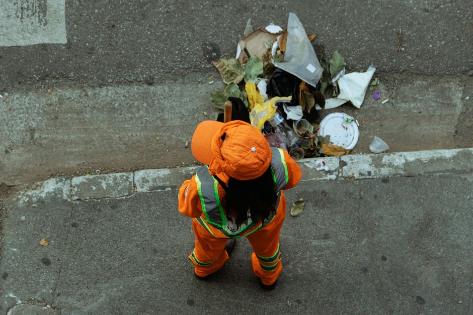 A person wearing a waterproof black jacket and hood, standing outdoors on a wet paved sidewalk during daytime, pushing a green trash cart lined with a black garbage bag. The scene features a street co